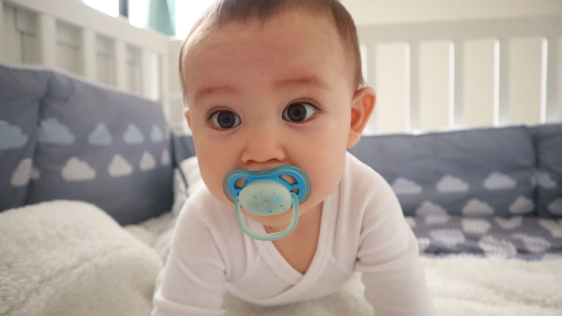 Baby sucking on a blue pacifier while lying on a bed.